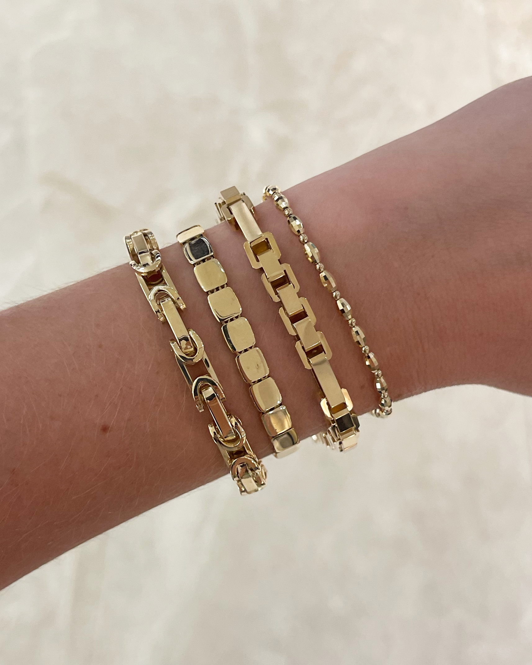 A close-up of a person's wrist wearing four gold-toned bracelets, including a curb link bracelet, a box link bracelet, a beaded chain bracelet, and a smaller chain bracelet, against a light beige marble background.