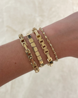 A close-up of a person's wrist wearing four gold-toned bracelets, including a curb link bracelet, a box link bracelet, a beaded chain bracelet, and a smaller chain bracelet, against a light beige marble background.