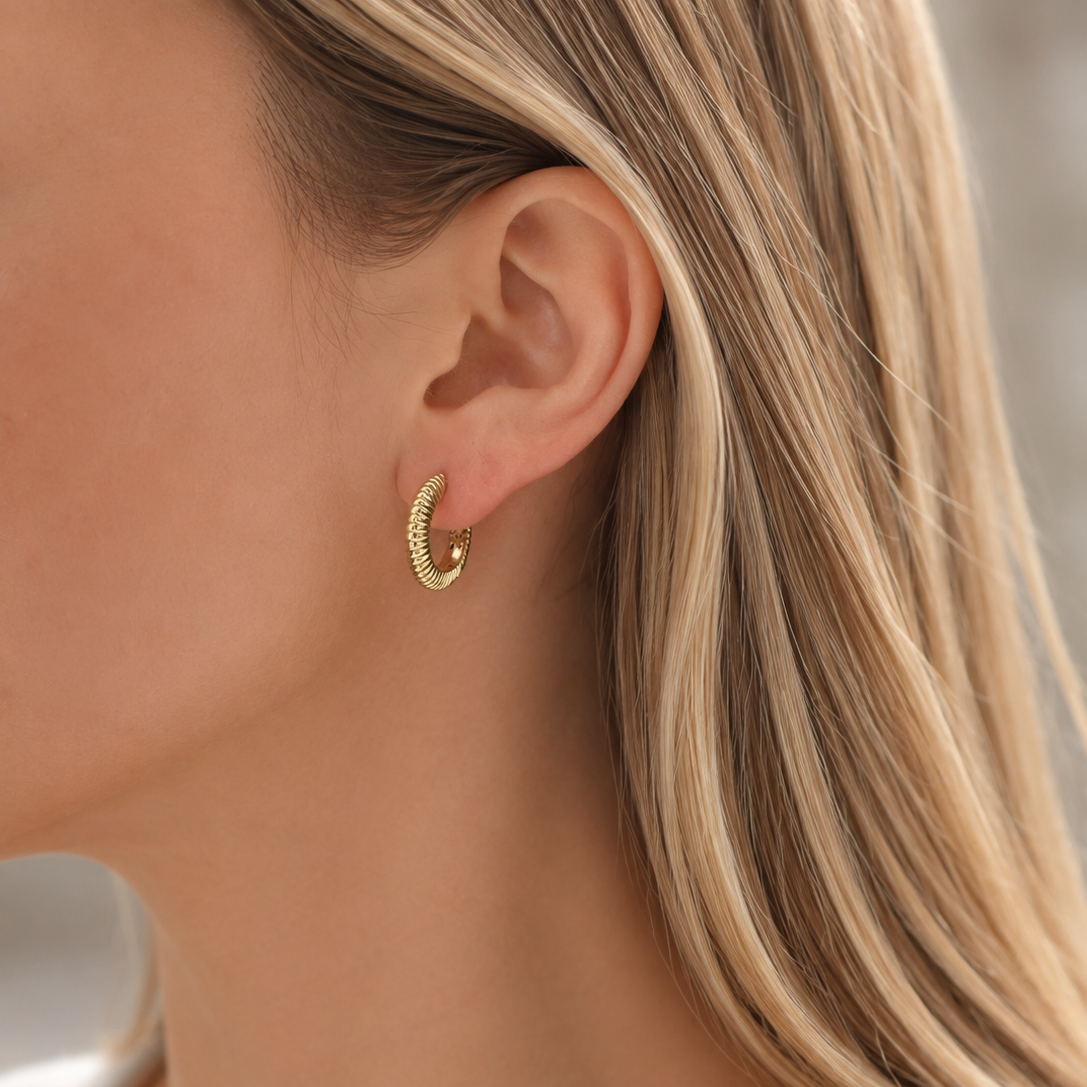 A close-up of a woman's ear wearing a small textured gold huggie hoop earring, with blonde hair visible against a soft neutral background.