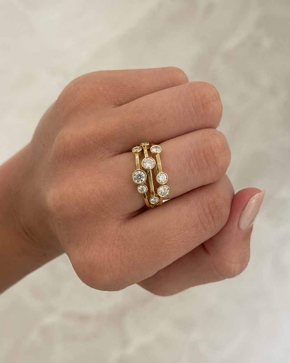 A close-up of a hand wearing a delicate gold ring set with small clear round gemstones, photographed against a light marble-like background.