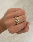 A close-up of a hand wearing a delicate gold ring set with small clear round gemstones, photographed against a light marble-like background.