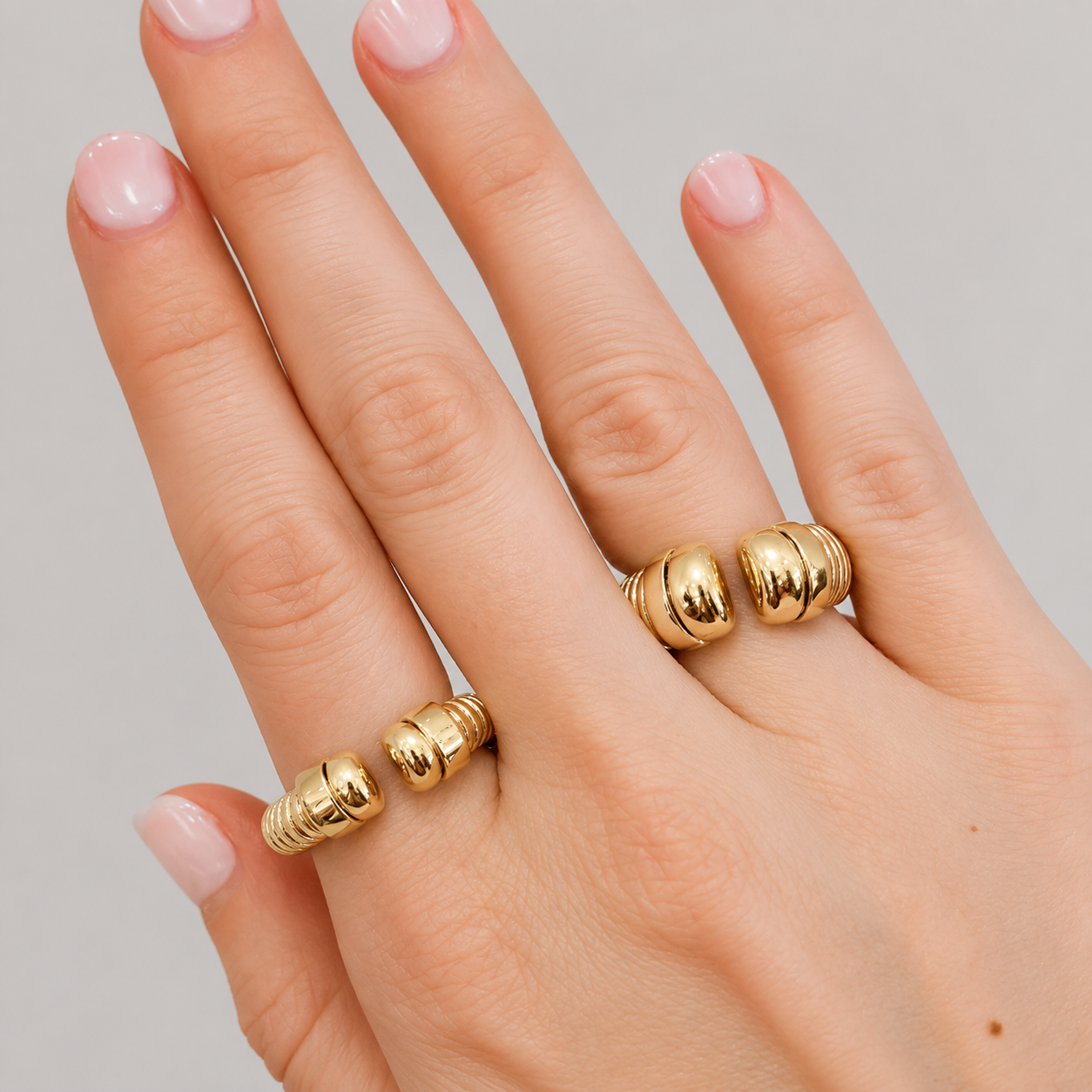A close-up of a hand wearing two gold rings with a textured, ribbed beaded design, set against a plain light gray background.