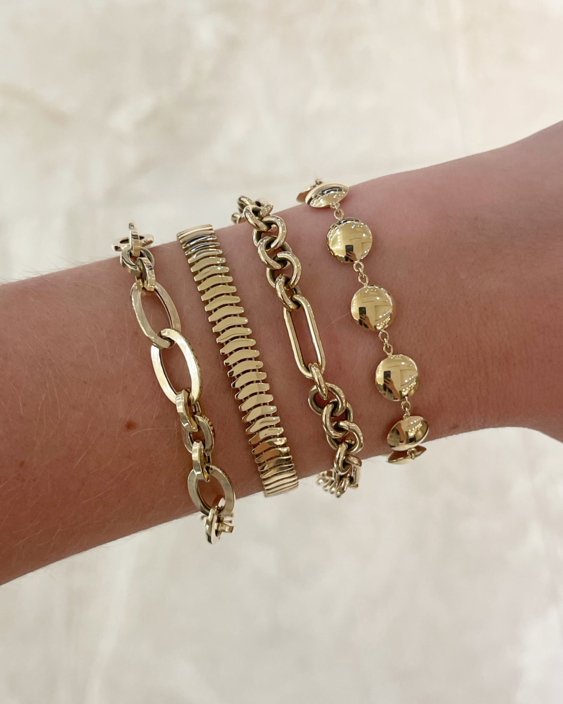 A close-up of a person's wrist wearing four gold-toned bracelets, including a snake chain, a link chain, a disc charm bracelet, and a textured chain.