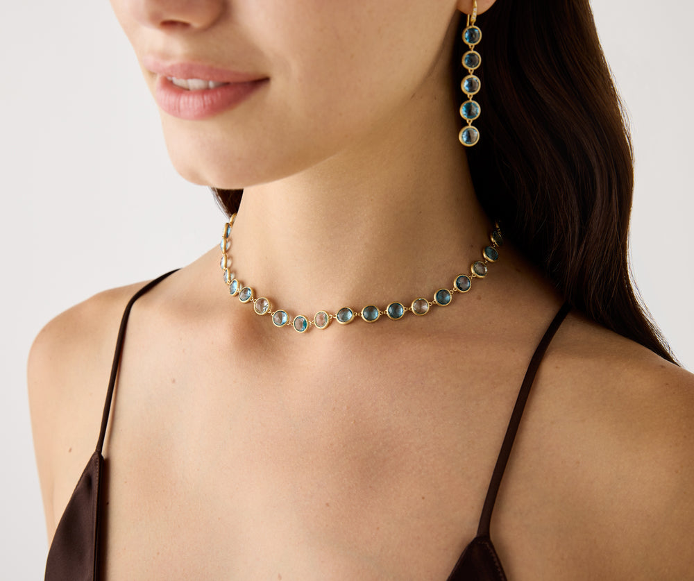 18K yellow gold station necklace featuring bezel-set, rose-cut round blue topaz gemstones arranged in a continuous line with a lobster clasp. Displayed on a model with matching earrings.