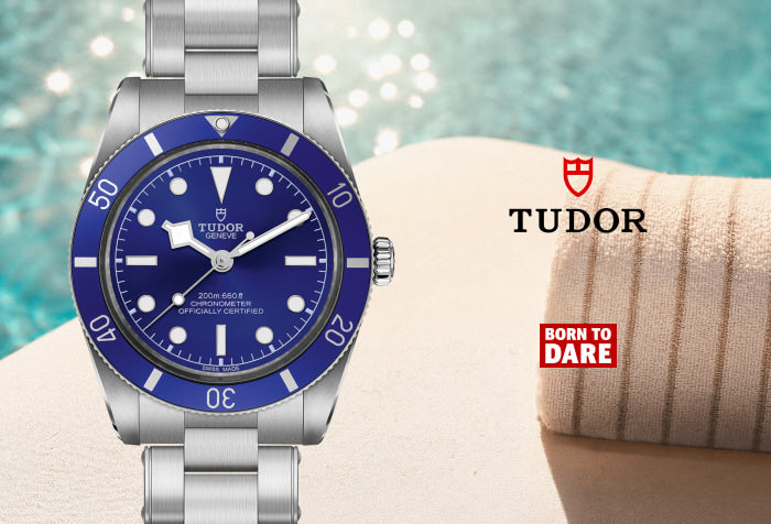 A Tudor watch with a silver stainless steel case and bracelet, featuring a vibrant blue dial and bezel. The watch is displayed on a light beige surface next to a rolled beige towel, with a blurred turquoise pool background.