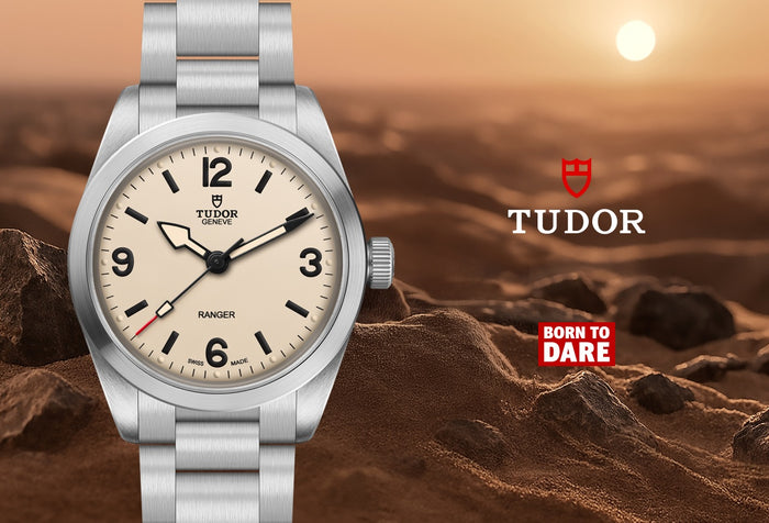 A Tudor Ranger wristwatch with a silver stainless steel bracelet and a cream-colored dial, set against a rocky desert landscape with a setting sun. The Tudor brand logo and 'Born to Dare' slogan are displayed on the right side of the image.